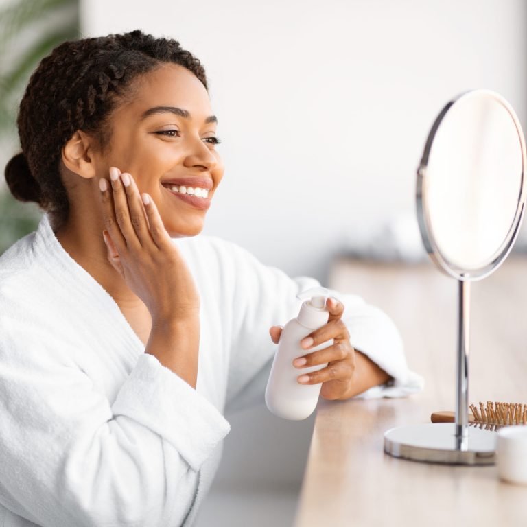 Smiling young black woman in white bathrobe applying skincare product, beautiful african american female looking at her reflection in table mirror in bright bathroom, doing beauty routine at home
