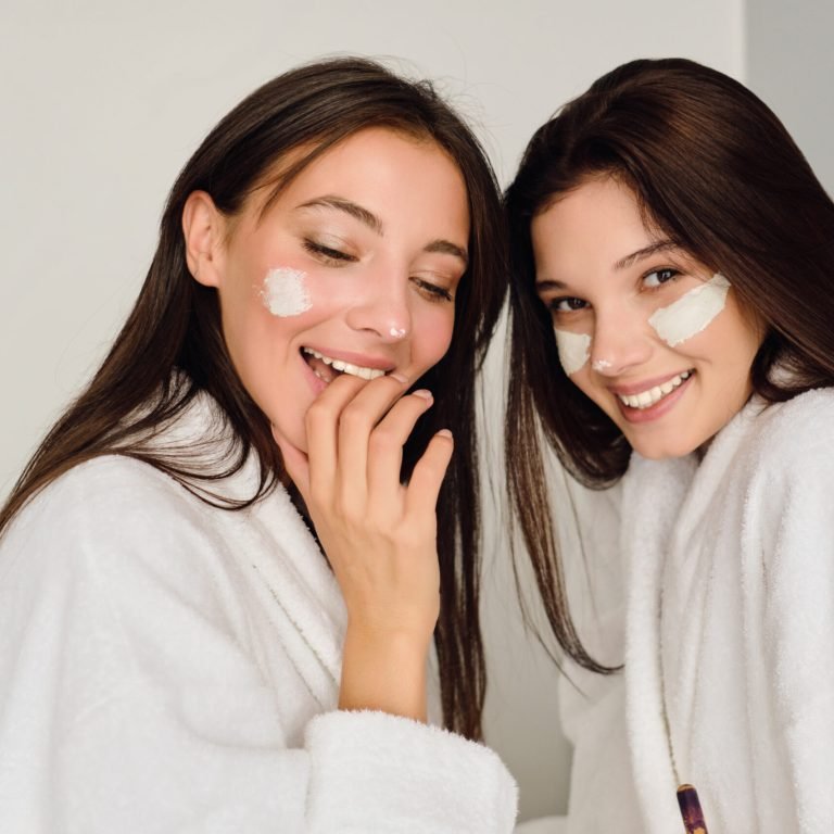 Two young beautiful women with dark hair in white bathrobes sitting on bed with cosmetic mask on faces happily looking in camera in modern hotel